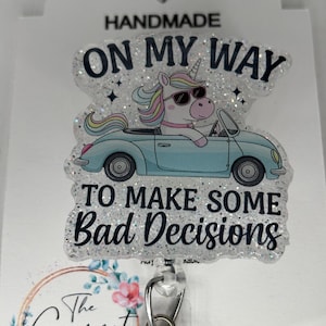 May include: A white glitter badge reel with a unicorn wearing sunglasses driving a blue car. The text on the reel reads "Handmade, On My Way To Make Some Bad Decisions". The reel is attached to a silver retractable badge holder with a clear plastic case. The reel is decorated with a floral design and the text "The Garnet Magnolia" and "@garnetmagnolia".
