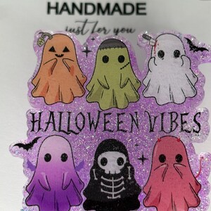 May include: A Halloween-themed badge reel with six cartoon ghosts in orange, green, white, purple, black, and pink. The text "HALLOWEEN VIBES" is displayed on a glittery purple background. The badge reel is labeled "HANDMADE just for you."