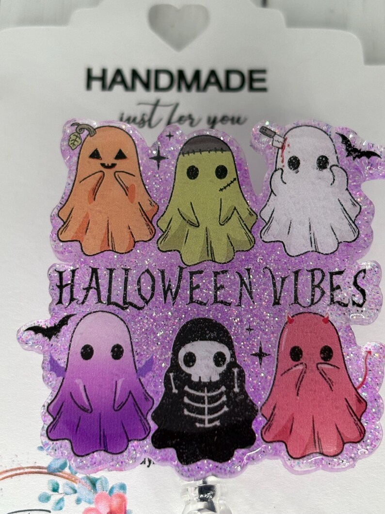 May include: A Halloween-themed badge reel with six cartoon ghosts in orange, green, white, purple, black, and pink. The text "HALLOWEEN VIBES" is displayed on a glittery purple background. The badge reel is labeled "HANDMADE just for you."