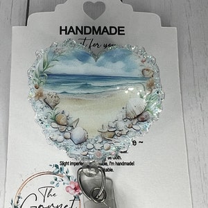 May include: A white card with a heart-shaped cutout and the text "HANDMADE" and "Just for you". A heart-shaped badge reel with a beach scene and seashells is attached to the card. The text "The Garnet Magnolia" is printed on the card.