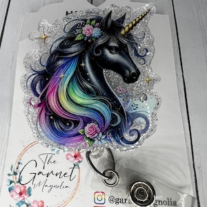 May include: A black glitter badge reel with a black unicorn with a rainbow mane and a gold horn. The unicorn has a floral crown and is surrounded by glitter. The badge reel has a clear plastic badge holder and a metal clip. The text "The Garnet Magnolia" is on the bottom of the reel.