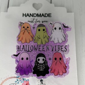 May include: A Halloween-themed badge reel featuring six cartoon ghosts in orange, green, white, purple, black, and pink. The text "HALLOWEEN VIBES" is displayed. The badge reel is attached to a white card with the text "HANDMADE just for you".