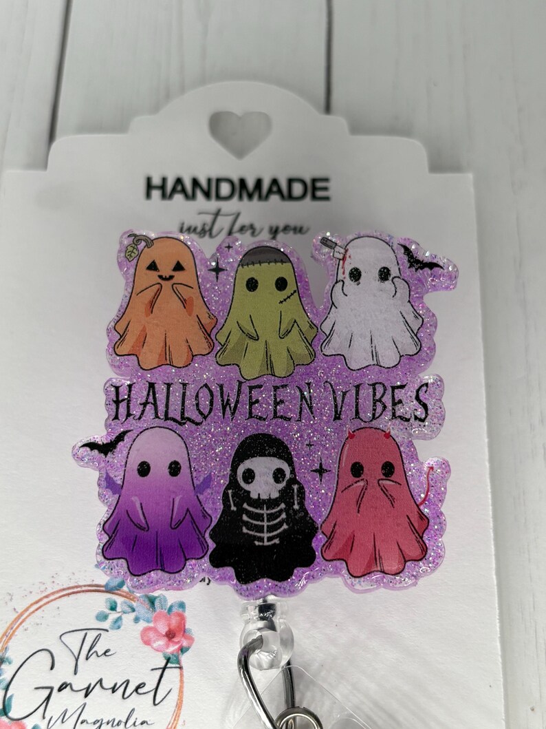 May include: A Halloween-themed badge reel featuring six cartoon ghosts in orange, green, white, purple, black, and pink. The text "HALLOWEEN VIBES" is displayed. The badge reel is attached to a white card with the text "HANDMADE just for you".