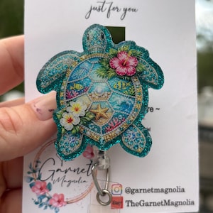 以下が含まれることがあります： 鮮やかな海のシーンとハイビスカスの花が描かれた、ウミガメの形をしたターコイズ色のグリッターバッジリール。「HANDMADE just for you」の文字とGarnet Magnoliaのロゴが入っています。