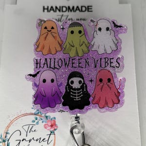May include: A Halloween-themed badge reel with six cartoon ghosts in orange, green, white, purple, black, and pink. The text "HALLOWEEN VIBES" is in purple. The badge reel is attached to a white card with the text "HANDMADE".