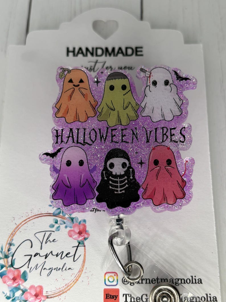 May include: A Halloween-themed badge reel with six cartoon ghosts in orange, green, white, purple, black, and pink. The text "HALLOWEEN VIBES" is in purple. The badge reel is attached to a white card with the text "HANDMADE".