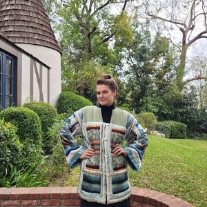 Puede incluir: Una mujer lleva una chaqueta larga de patchwork multicolor con cierre de botones. La chaqueta tiene una variedad de telas y patrones, incluyendo diseños florales, geométricos y a rayas. La mujer está de pie frente a una pared de ladrillo y un césped verde.