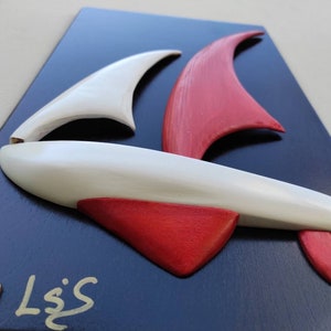 Könnte beinhalten: Eine weiße und rote Holzskulptur eines Segelboots, montiert auf einem dunkelblauen Hintergrund. Die Skulptur trägt den Titel "L.S.S."