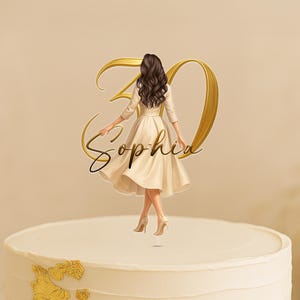Op de afbeelding: Taarttopper met een vrouw in een crèmekleurige jurk en het nummer 30 in goud. De naam "Sophia" is cursief geschreven. De taart is wit met gouden details.