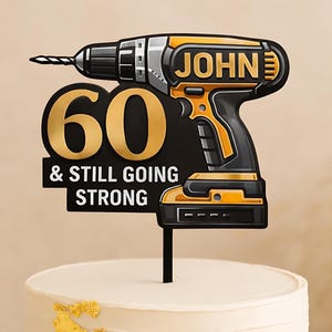 Peut inclure: Décoration de gâteau en forme de perceuse noire, jaune et argentée, avec le nom "JOHN" et le texte "60 & STILL GOING STRONG". Le gâteau est blanc avec des décorations dorées. Thème anniversaire.