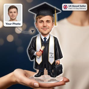 May include: A graduation caricature figurine of a man in a black cap and gown, holding a diploma and briefcase. The figurine has a cartoon-like appearance and features the text "Class of 2026". The figurine is held in a person's hands.