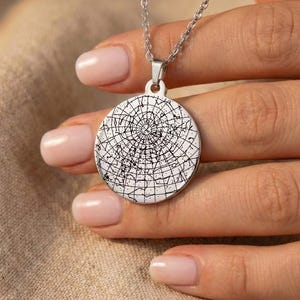 Puede incluir: Un collar con colgante plateado con un dije circular con un diseño geométrico en blanco y negro. El collar se sostiene en una mano con uñas cuidadas, mostrando el intrincado patrón y el brillo metálico del colgante.