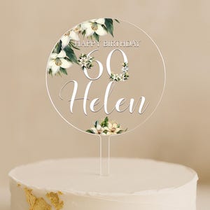 Può includere: Un cake topper trasparente con le parole "Happy Birthday 60 Helen" in corsivo bianco. Il design include elementi floreali con fiori bianchi e foglie verdi, perfetto per un 60° compleanno.