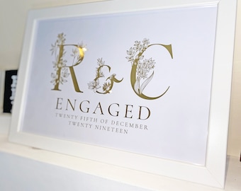 May include: A white framed print with gold lettering announcing an engagement. The print features the initials "R & C" with floral designs and the text "ENGAGED TWENTY FIFTH OF DECEMBER TWENTY NINETEEN".
