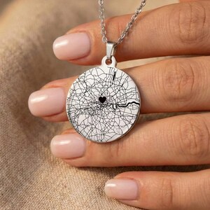 Puede incluir: Collar con colgante plateado con un colgante circular que muestra un diseño de mapa en blanco y negro. Un corazón negro marca una ubicación en el mapa. El collar lo sostiene una persona.