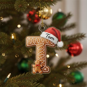 May include: A clear Christmas tree decoration in the shape of the letter "T", decorated with a gingerbread man and a Santa hat. The ornament is brown with white icing details and the name "Tom" is written on the hat. It hangs from a gold string.