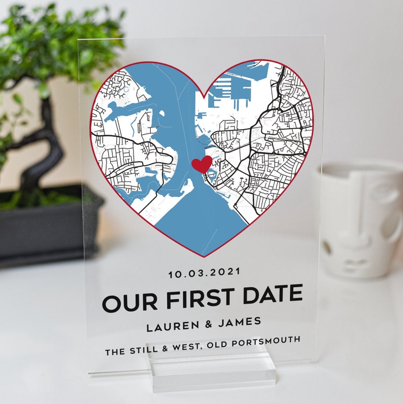 May include: A clear acrylic sign with a red heart-shaped map of a city. The map shows streets and a body of water. The date 10.03.2021, the text "OUR FIRST DATE", the names "LAUREN & JAMES", and the location "THE STILL & WEST, OLD PORTSMOUTH" are printed on the sign.