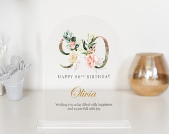 May include: A clear acrylic sign with a gold and floral design. The number 90 is displayed with a floral wreath. The text "HAPPY 90TH BIRTHDAY Olivia Wishing you a day filled with happiness and a year full with joy" is printed on the sign.