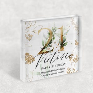 Puede incluir: Un bloque acrílico cuadrado blanco con un diseño floral dorado y verde. El número 21 está en dorado con hojas y ramas verdes. El texto "Victoria Feliz cumpleaños Feliz cumpleaños Victoria, te queremos mucho!" está en negro.