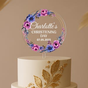 Może przedstawiać: Przezroczysty topper na tort z akrylu z kwiatowym wieńcem w odcieniach fioletu i niebieskiego. Tekst brzmi "Charlotte's CHRISTENING DAY 07.05.2025". Umieszczony na białym torcie ze złotymi zdobieniami.