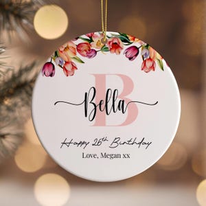 Peut inclure: Une décoration de Noël en céramique blanche avec un motif floral rose et le nom "Bella" en lettres roses. La décoration comprend également le texte "Happy 26th Birthday" et "Love, Megan xx".
