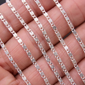 May include: A close-up of a silver chain with a delicate, patterned design. The chain is shown in multiple lengths, laying on a hand.