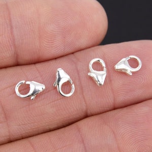 May include: Four silver lobster claw clasps, a type of jewelry finding, are shown on a hand. The clasps are small and have a shiny finish.