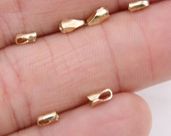 May include: Close-up of several small, gold-colored jewelry findings. The findings are various shapes, including tubes and teardrop shapes, with small loops. The items are on a light-colored background.