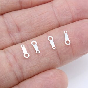 May include: Four small, silver-colored jewelry connectors with a rectangular body and a circular loop at one end. The connectors are displayed on a light-colored background.