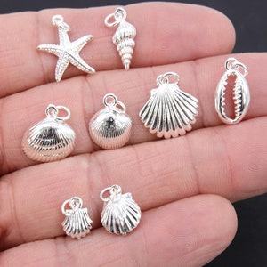 Puede incluir: Una colección de colgantes de plata con forma de conchas marinas y una estrella de mar. Los colgantes se muestran en una mano.