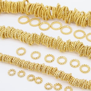 May include: Gold-toned twisted wire jump rings in various sizes. The jump rings are arranged in a scattered pattern on a white background. A Girls Gems logo is in the bottom right corner.