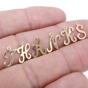 May include: Gold-toned metal letter charms spelling out the word "THANKS". The charms are in a cursive font and are arranged in a line on a hand.