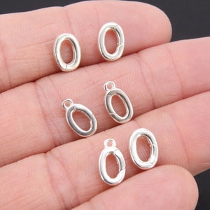 May include: Six silver oval-shaped jump rings with a loop on one end. The jump rings are on a hand.