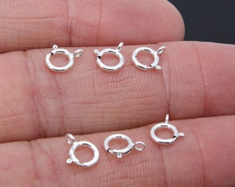 May include: Six silver-toned jump rings, also known as lobster clasps, are displayed on a hand. The jump rings are approximately 1/2 inch in diameter.
