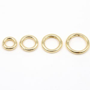 May include: Four gold-colored, circular jewelry clasps of varying sizes are arranged horizontally against a white background. The clasps appear to be made of metal and have a shiny finish. The smallest clasp is on the left, with the sizes increasing to the right.