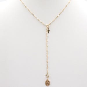May include: A gold-colored necklace with a cross pendant and a small medallion. The necklace is displayed on a white mannequin. The medallion has a religious image. The necklace is delicate and has a Y-shaped design.