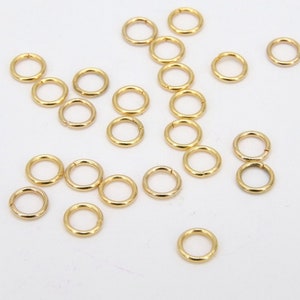 Puede incluir: Una gran cantidad de anillos de salto dorados, esparcidos sobre una superficie blanca. Estos anillos se utilizan comúnmente en la fabricación de joyas.