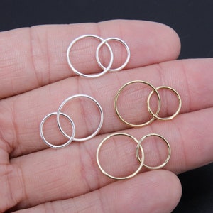 May include: A set of three pairs of hoop earrings, two pairs are silver and one pair is gold. The earrings are arranged in a hand, with the larger hoops on top and the smaller hoops on the bottom.