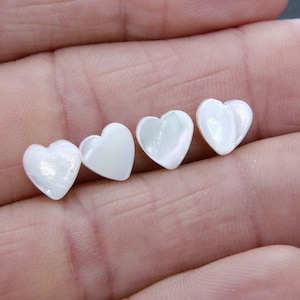 May include: Four white heart-shaped mother of pearl cabochons. The cabochons are arranged in a row from left to right, with the largest cabochon on the right and the smallest on the left.