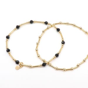 May include: Two gold-colored bracelets with black beads. The bracelets have a delicate design, with small gold beads and longer gold bars. A small gold charm hangs from one bracelet. The background is white.