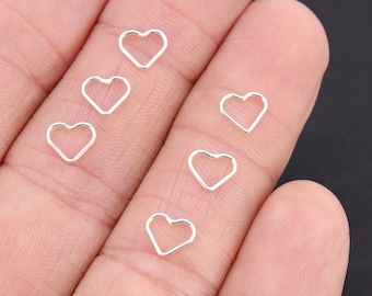 May include: A hand holding six silver heart-shaped jump rings. The rings are open and ready to be used for jewelry making.