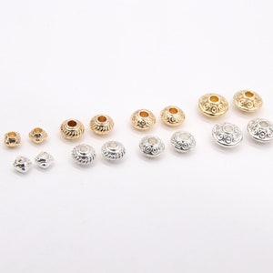 May include: Assortment of gold and silver metal beads with a decorative design. The beads have a round shape with a hole in the center.