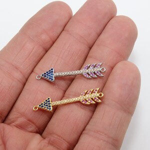 May include: Two gold arrow charms with crystal embellishments. One arrow charm has blue crystals and the other has pink crystals.
