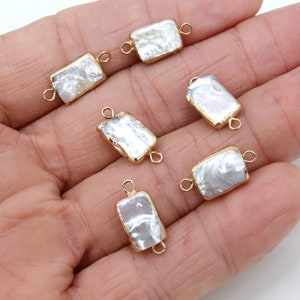 May include: Six white rectangular pearl connectors with gold-toned jump rings. The connectors are on a hand.