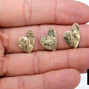 May include: Three gold-colored heart-shaped charms with a hammered texture. The charms have a hole at the top for attaching to jewelry or other crafts.