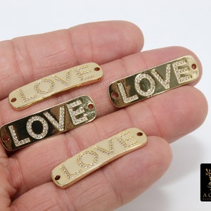 May include: Four gold-colored metal charms with the word "LOVE" spelled out in clear rhinestones. The charms have two holes for attaching to jewelry.