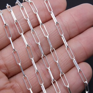 Pode incluir: Um colar de corrente de prata com um estilo de elo que se assemelha a um clipe de papel. A corrente é mostrada em várias fitas, sobre uma mão.