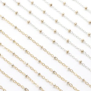 May include: Multiple delicate satellite chains arranged in parallel rows. The collection includes silver-toned chains with small silver beads and gold-toned chains with small gold beads. A small black logo tag with gold text is in the bottom corner.