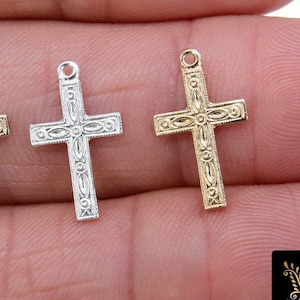 May include: Four different colored cross charms, silver, gold, and two shades of gold, with intricate designs. The charms are displayed on a hand.