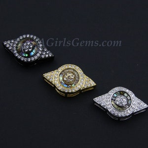 May include: Three eye-shaped charms with a round abalone shell center surrounded by clear rhinestones. The charms are in black, gold, and silver.